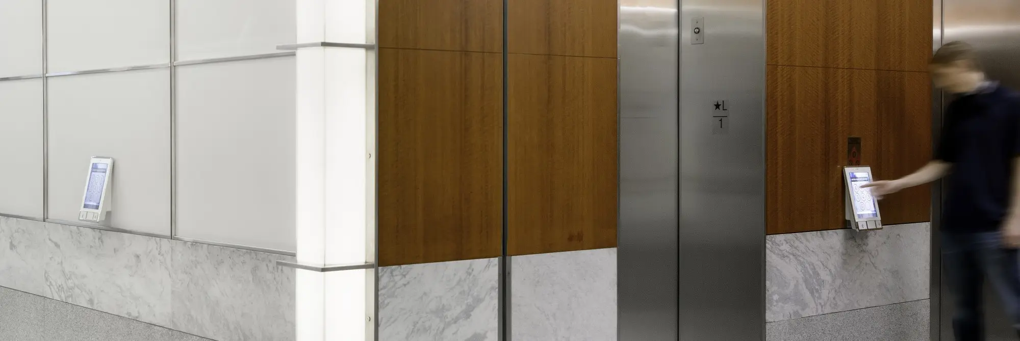 A series of elevators from the side with a man using a destination dispatch kiosk.